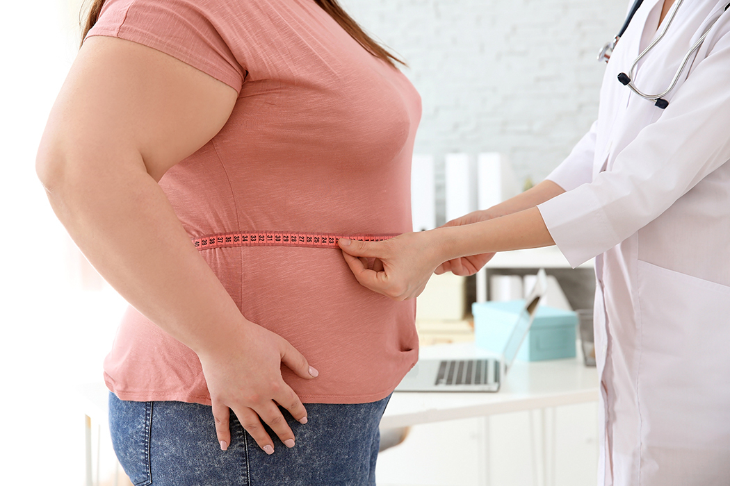 A doctor in a white coat uses a tape measure to take an overweight woman’s waist measurement in an office.